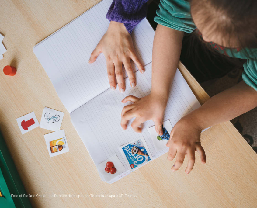 Foto con due persone, una bambina e una logopedista, durante l'attività di logopedia. Sono mostrate solo le mani che incollano su un quaderno varie immagini.