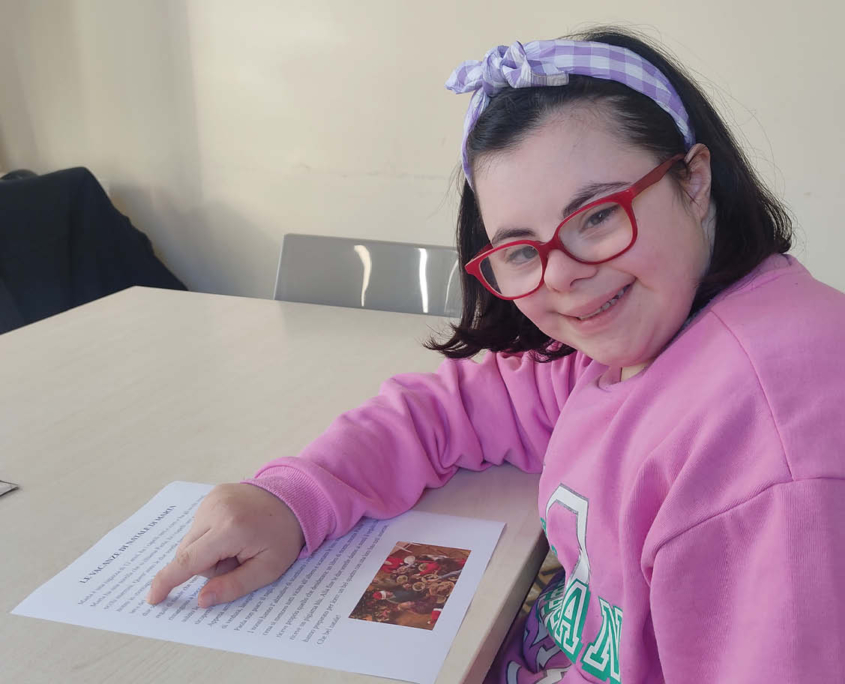 Una bambina vestita di rosa, sorride mentre tiene il segno di un testo che sta leggendo, durante l'attività di Logopedia.